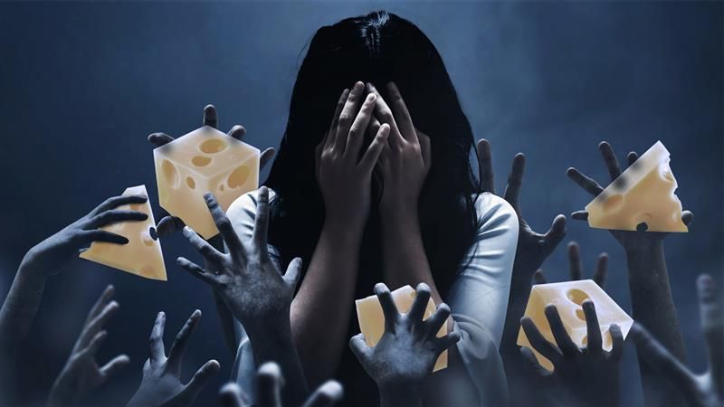 The image shows a woman with long black hair standing in a dark almost misty room. She is covering her face with her hands like she is in despair. Around her, numerous hands are reaching up towards her and they have been digitally edited so they hold pieces of cheese. 