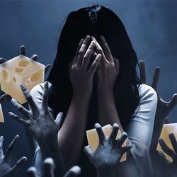 The image shows a woman with long black hair standing in a dark almost misty room. She is covering her face with her hands like she is in despair. Around her, numerous hands are reaching up towards her and they have been digitally edited so they hold pieces of cheese. 