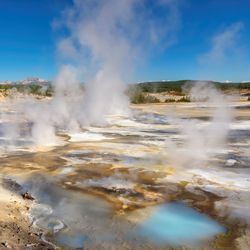 Geysers in Norris Basin, Yellowstone National Park, Wyoming, USA. The landscape is rocky with a number of pools that appear bright turquoise; there are clouds of steam rising, and in the distance green plants cover the terrain.