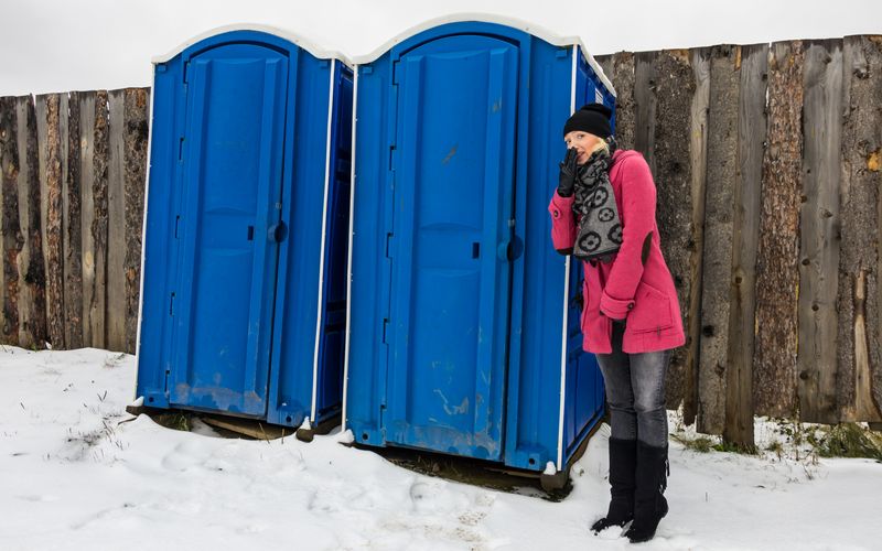 woman wearing a pink coat and black hat, scarf and gloves waiting by two blue portapotties outside in the snow