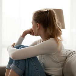 Side view young woman looking away at window sitting on couch at home. 